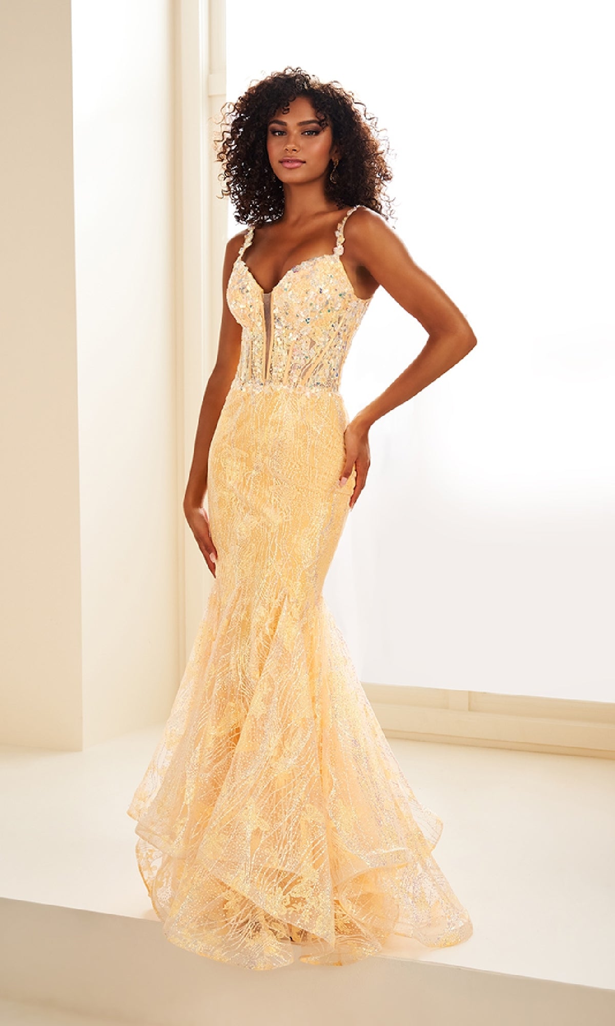 A woman stands in a bright room wearing the Ellie Wilde EW36038 Glow-in-the-Dark Prom Dress, a floor-length yellow lace gown with thin straps, a structured bodice, beaded details, and sparkling sequin accents.